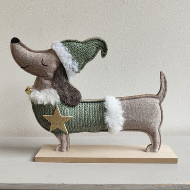Christmas sausage dog / dachshund wearing a green sweater with a gold star and a festive hat on a light background. By Heaven Sends