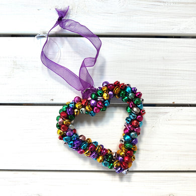Colorful heart-shaped bell ornament with a purple ribbon on a white wooden background.  Fair Trade by Namaste