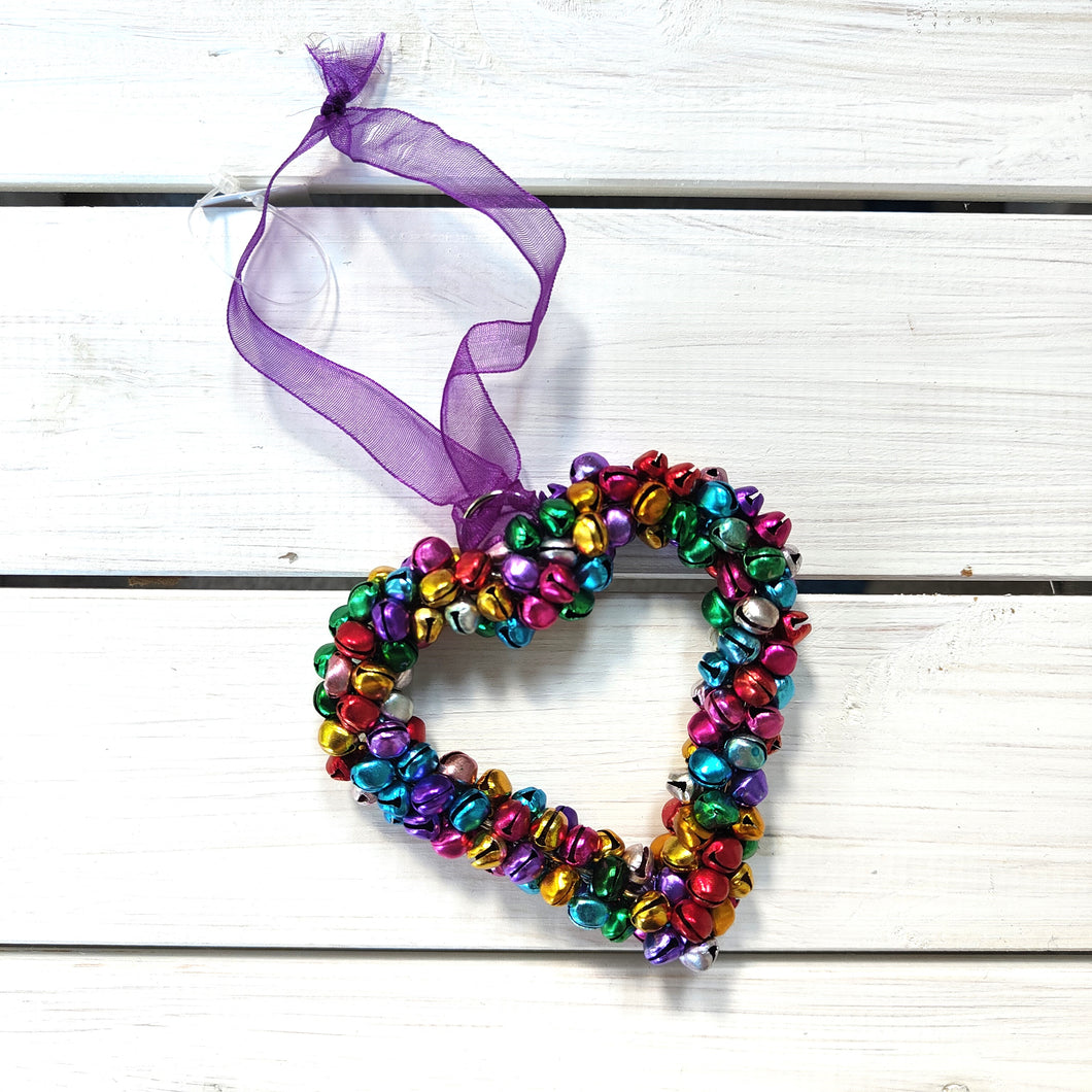 Colorful heart-shaped bell ornament with a purple ribbon on a white wooden background.  Fair Trade by Namaste
